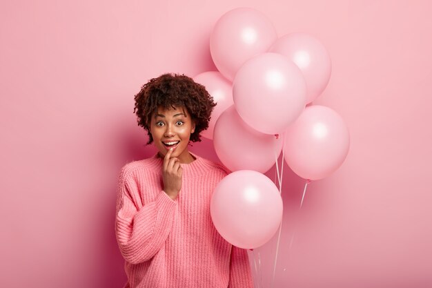 Foto interna de um jovem afro-americano feliz e otimista surpreso por estar presente, segura balões de ar, usa um macacão enorme, mantém o dedo no lábio inferior, satisfeito por estar presente na celebração da festa.