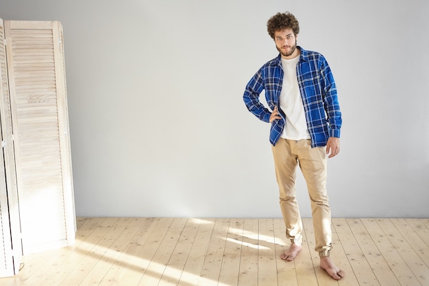 Foto interna de um jovem modelo europeu barbudo e atraente, vestindo jeans bege da moda e camisa azul xadrez, posando descalço no chão de madeira em casa, mantendo a mão na cintura
