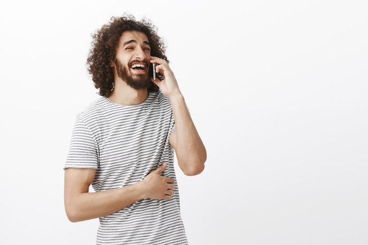 Foto grátis foto interna de um namorado atraente alegre com barba e penteado afro, olhando para o lado enquanto fala no smartphone, rindo alto