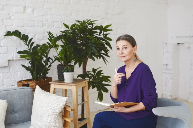 Foto interna de uma atraente treinadora de negócios europeia de meia-idade, pensativa, sentada em uma sala espaçosa e iluminada, rodeada de móveis modernos e vasos de plantas, verificando a agenda em seu diário