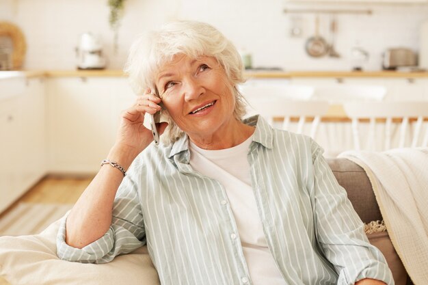 Foto interna de uma encantadora mulher de cabelos grisalhos sênior, segurando um telefone inteligente genérico perto do ouvido, tendo problemas de audição, conversando com a amiga, sentada confortavelmente no sofá da sala de estar