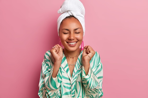 Foto interna de uma feliz mulher de pele escura em pé com os olhos fechados, cerrando os punhos, alegra-se com as boas notícias, vestida com manto doméstico, toalha de banho embrulhada. Dona de casa passa fim de semana em casa. Atmosfera aconchegante