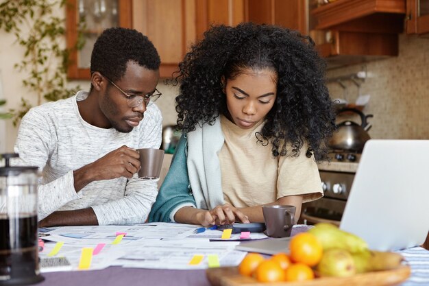 Foto interna de uma garota africana séria usando calculadora enquanto paga contas, sentada à mesa da cozinha