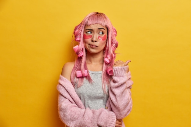 Foto interna de uma garota asiática confusa com longos cabelos rosa, faz penteado encaracolado com rolos, aponta para o espaço da cópia e tem uma expressão desagradada