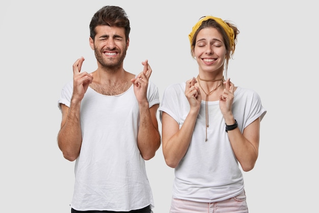 Foto grátis foto interna de uma jovem caucasiana supersticiosa e um homem com os dedos cruzados, tenha esperança, sonhe com algo agradável, fique perto um do outro, feche os olhos, sorria amplamente, acredite na boa sorte