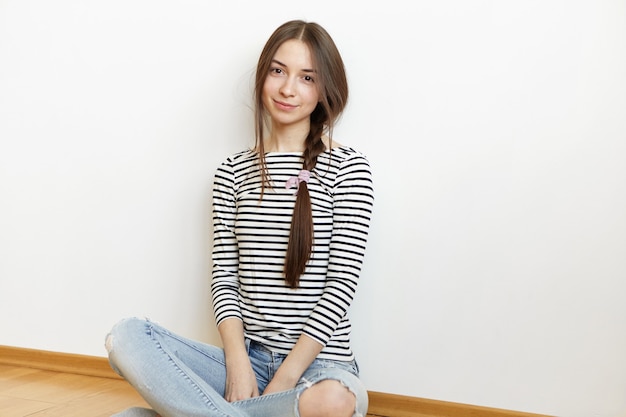 Foto interna de uma jovem encantadora com uma trança de cabelo bagunçado relaxando em casa depois da faculdade