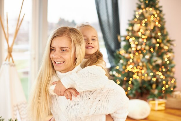 Foto interna de uma jovem feliz com cabelo comprido dando carona para sua adorável filhinha, se divertindo e brincando na sala de estar com uma árvore de Natal decorada e brilhante