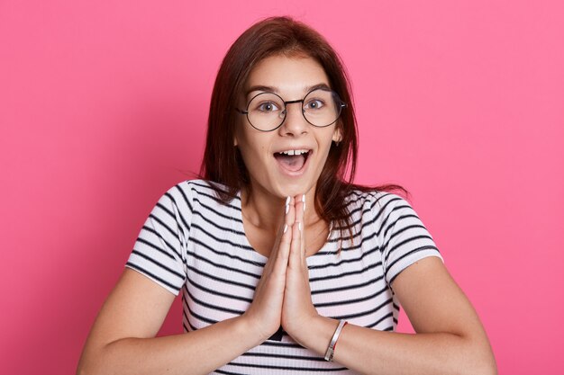 Foto interna de uma linda jovem fêmea com expressão positiva, mantém as palmas das mãos juntas, tem uma expressão surpresa, posa contra uma parede rosada, mantendo a boca bem aberta.