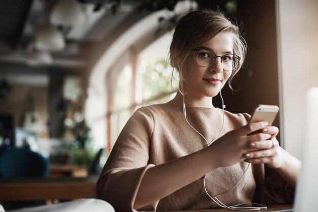 Foto interna de uma linda mulher caucasiana, elegante e feliz, com cabelos louros dentro de óculos, segurando um smartphone e usando fones de ouvido enquanto assiste ao vídeo, o que distrai ao olhar e sorrir