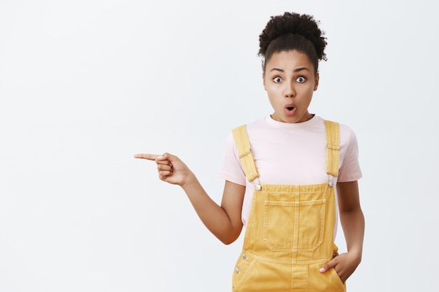 Foto interna de uma mulher afro-americana expressiva e bonita impressionada de macacão amarelo, deixando cair o queixo de espanto, dobrando os lábios e apontando para o espaço da cópia à esquerda sobre a parede cinza