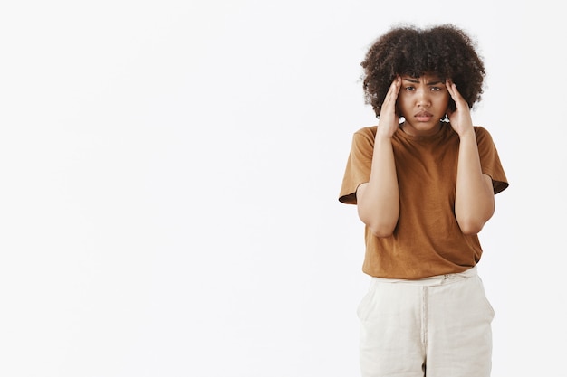 Foto interna de uma mulher afro-americana intensa e incomodada com um penteado afro encaracolado de mãos dadas nas têmporas, franzindo a testa e olhando com uma expressão doente e cansada tentando se concentrar