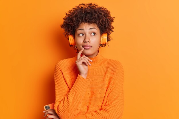 Foto interna de uma mulher afro-americana pensativa, concentrada acima, mantém o dedo no queixo e considera algo em mente enquanto ouve uma melodia calma em fones de ouvido estéreo