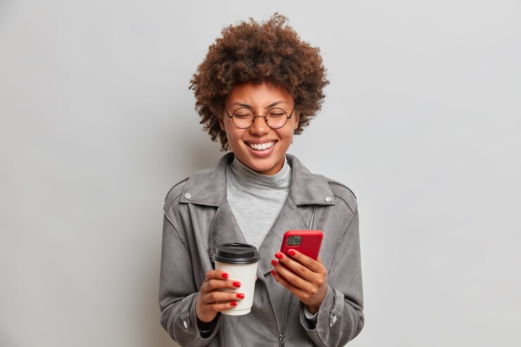Foto grátis foto interna de uma mulher alegre e otimista que começa a rir, bebe café para viagem, segura o telefone celular, espera por alguém, caminha durante o dia de folga, expressa emoções positivas