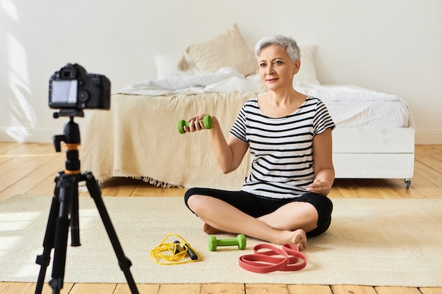 Foto interna de uma mulher aposentada saudável em roupas elegantes, sentada no chão do quarto em frente ao tripé da câmera, gravando um vídeo tutorial de fitness para idosos, mostrando exercícios com halteres