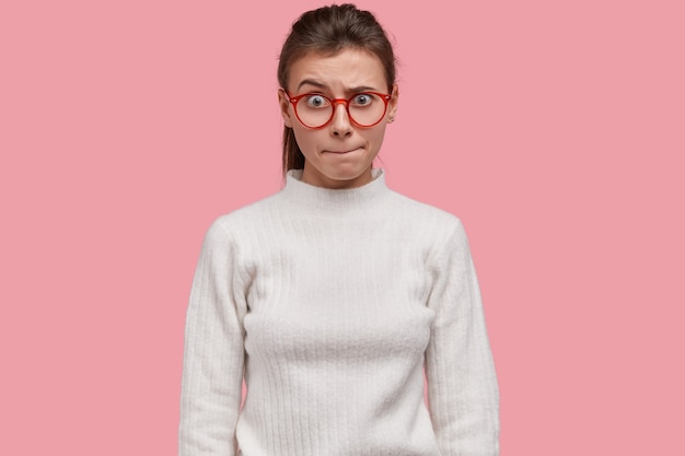 Foto interna de uma mulher bonita pressionando os lábios, levantando as sobrancelhas em perplexidade, reagindo a algo inesperado, vestindo um suéter branco