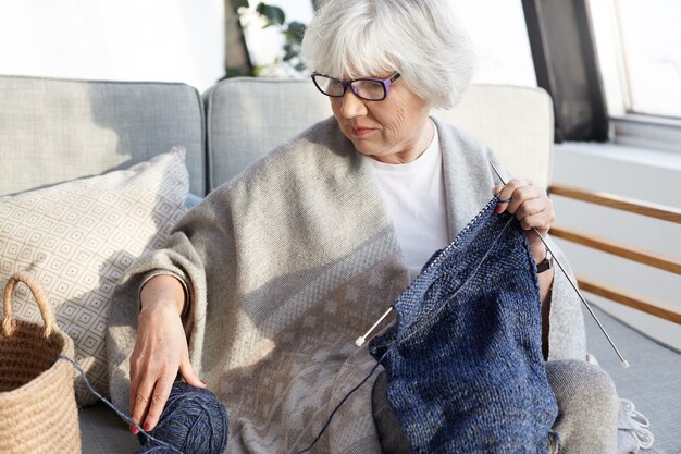 Foto interna de uma mulher idosa séria e concentrada, com cabelos grisalhos, sentada no sofá da sala de estar usando óculos, tricotando roupas quentes de inverno para seu site na Internet, vendendo produtos caseiros online