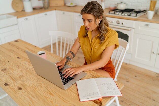 Foto interna de uma mulher loira sorridente usando laptop durante o café da manhã em sua cozinha moderna e leve.