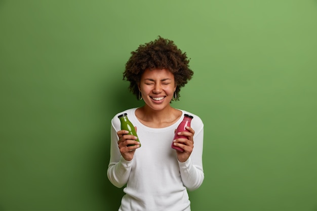 Foto interna de uma mulher sorridente muito feliz segurando garrafas de espinafre e smoothie de morango, gosta de beber bebida desintoxicante, posa com uma bebida fresca preparada na hora, isolada no fundo verde