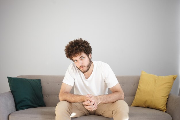 Foto interna do modelo masculino sério bonito jovem com barba por fazer em uma camiseta branca e jeans bege, posando na acolhedora sala de estar moderna na parede branca, sentado no sofá, apertando as mãos e