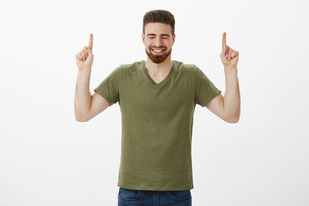 Foto interna do namorado barbudo bonito muito feliz torcendo e se sentindo alegre, sorrindo e rindo com os olhos fechados, apontando com o dedo indicador levantado para cima contra a parede branca