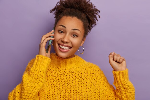 Foto isolada de adolescente feliz e alegre aprecia uma conversa, telefones via celular, sorri amplamente, levanta o braço, vestida com roupas de inverno