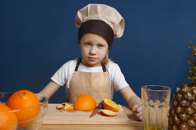 Foto isolada de chef infantil sério do sexo masculino com olhos azuis e cabelo loiro fazendo frutas frescas ou salada