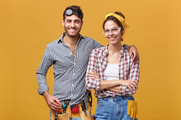 Foto isolada de jovens funcionários europeus confiantes e atraentes de manutenção, vestidos com macacões e roupas de proteção, equipados com instrumentos, prontos para o trabalho, com sorrisos felizes