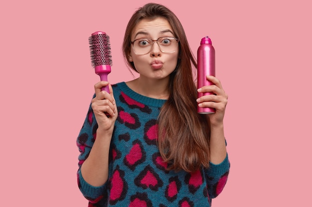 Foto isolada de mulher bonita dando mwah, dobra os lábios, tem cabelo comprido e cuidado, segura o pente e spray para cabelo, usa grandes óculos ópticos