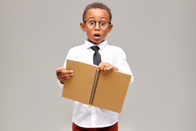 Foto isolada de um aluno africano chocado emocionalmente, vestindo camisa branca, gravata preta e óculos com olhar surpreso e surpreso, mantendo a boca aberta, segurando um caderno em branco aberto nas mãos