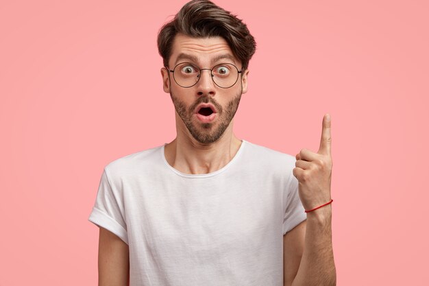 Foto isolada de um homem surpreso com a barba por fazer engasga de espanto, aponta com o dedo da frente para cima, usa óculos redondos e camiseta branca, modelos contra uma parede rosa, demonstra algo acima