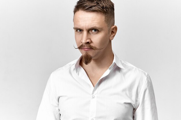 Foto isolada de um jovem empresário bem-sucedido com bigode e cavanhaque posando no estúdio, vestindo uma camisa formal branca, olhando para a câmera com um sorriso confiante