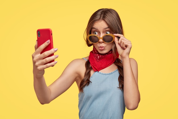Foto isolada de uma adolescente na moda faz beicinho, olha através de óculos escuros da moda, usa bandana vermelha perto do pescoço, segura o telefone inteligente, faz um retrato de selfie, aproveita o tempo livre, fica de pé sobre a parede amarela
