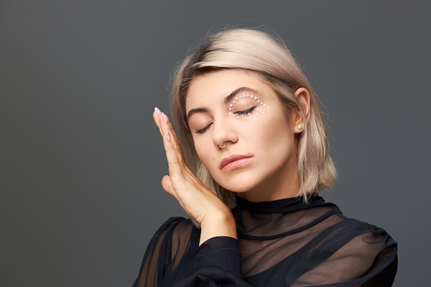 Foto isolada de uma adorável jovem de 20 anos com uma blusa preta transparente, mantendo os olhos fechados e tocando suavemente sua pele macia usando um piercing no nariz, corte de cabelo estiloso e maquiagem brilhante