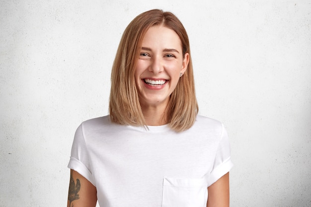 Foto isolada de uma jovem alegre e satisfeita com uma aparência agradável, sorriso largo e encantador, braço tatuado, vestida com uma camiseta branca casual, poses em estúdio, feliz por alcançar o sucesso no trabalho e na vida