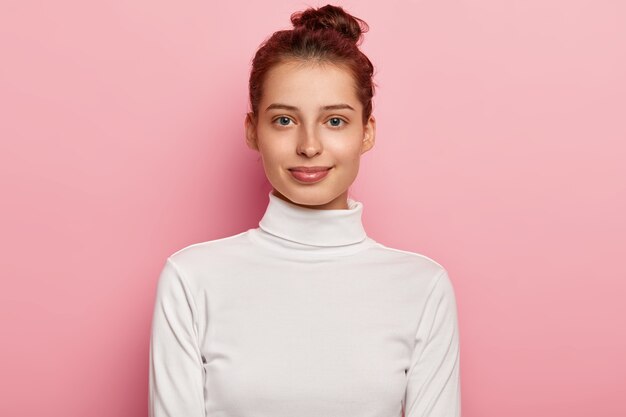 Foto isolada de uma jovem de aparência agradável usa gola alta branca, tem coque de cabelo, parece com expressões faciais calmas, poses contra um fundo rosa.