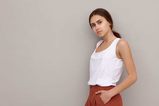 Foto isolada de uma jovem e atraente modelo feminina caucasiana, vestida com roupas elegantes, encostada na parede em branco com copyspace para seu texto ou informações publicitárias