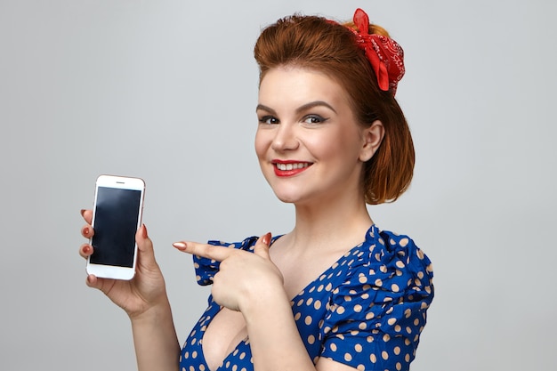 Foto grátis foto isolada de uma jovem e elegante modelo feminina vestindo roupas retrô e batom vermelho, sorrindo alegremente, promovendo um aparelho eletrônico moderno, segurando um celular genérico