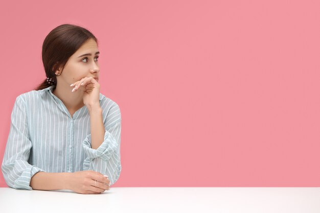 Foto isolada de uma jovem empresária atraente vestindo uma camisa listrada azul, sentada em sua mesa de escritório com a mão no queixo, olhando pensativamente na parede rosa em branco com copyspace para sua informação