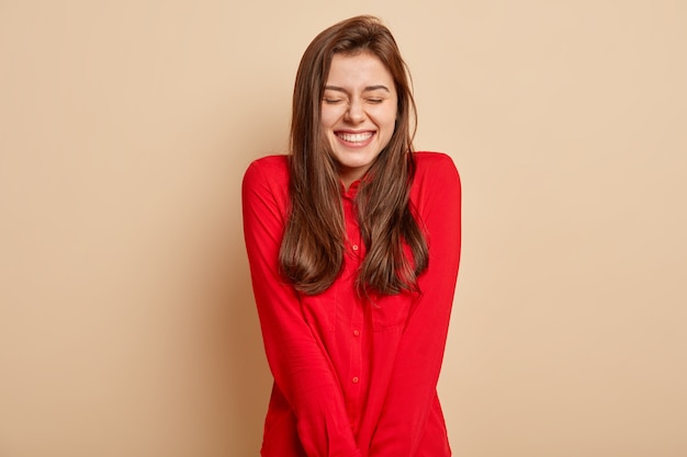 Foto isolada de uma jovem europeia positiva sente prazer e prazer, mantém os olhos fechados, sorri amplamente, vestida com roupas vermelhas, isolada sobre a parede marrom. Pessoas, conceito de emoções