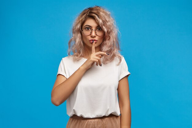 Foto isolada de uma linda aluna com cabelo rosado cobrindo os lábios com o dedo da frente, pedindo silêncio, preparando-se para os exames