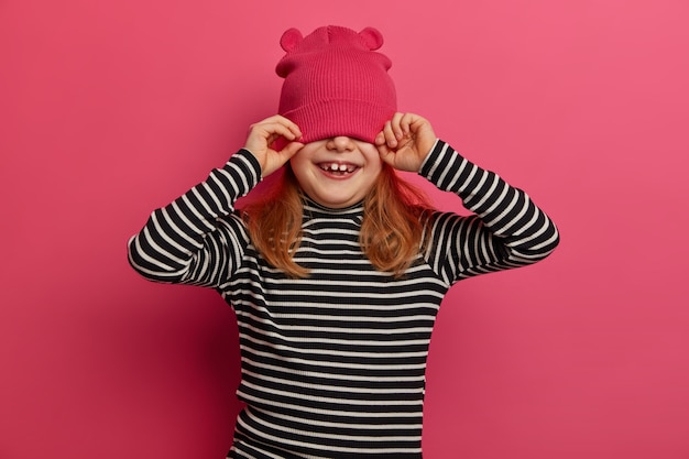 Foto isolada de uma linda menina de quatro anos usa um macacão listrado e um chapéu rosa, se diverte e cobre os olhos, gosta de passar o tempo no círculo familiar, isolado na parede rosa. crianças, emoções, roupas