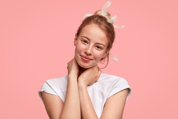 Foto isolada de uma linda mulher com pele saudável, mantém as mãos no pescoço, sorri positivamente, tem uma aparência atraente, penas no cabelo, usa uma camiseta branca casual, ouve algo bom. Conceito de dormir