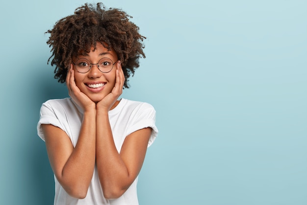 Foto grátis foto isolada de uma mulher afro com aparência positiva segura as duas mãos no rosto, sorri feliz, alegra-se por passar um bom fim de semana com o namorado, isolada sobre uma parede azul, espaço vazio para seu anúncio