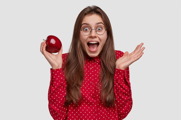 Foto isolada de uma mulher atraente com uma expressão alegre, segurando uma maçã vermelha com uma das mãos, expressando sentimentos positivos, usando uma camisa formal