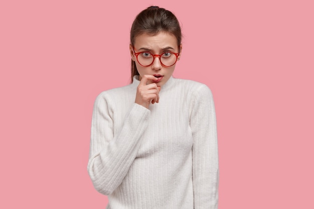 Foto isolada de uma mulher séria de aparência agradável mantendo o dedo da frente perto dos lábios, olhando seriamente através dos óculos, vestida com roupas casuais brancas