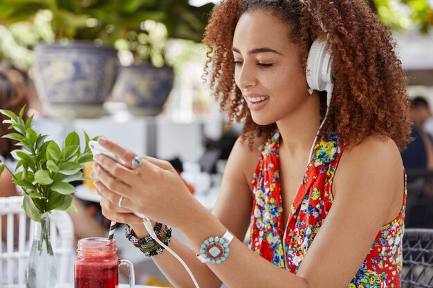 Foto lateral de linda jovem encantada se diverte e ri enquanto ouve a composição favorita em fones de ouvido, digita mensagem para o namorado