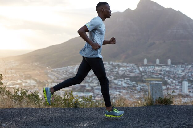 Foto lateral de um homem bonito e ativo correndo contra belas paisagens montanhosas, fotografado em movimento, gosta de se exercitar, é muito rápido e enérgico, usa roupas esportivas. Atleta negro do lado de fora