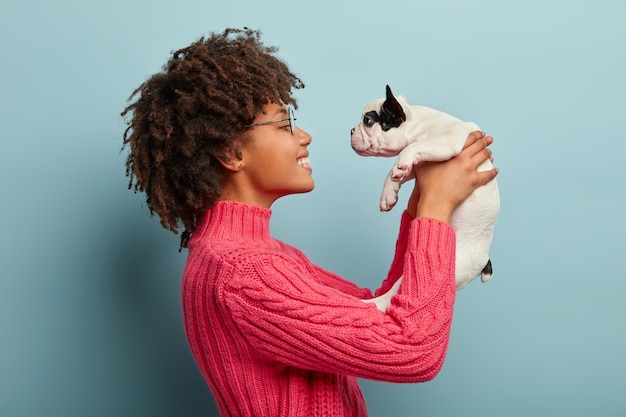 Foto grátis foto lateral de uma fêmea afro positiva brincalhona cuida de um cachorrinho com pedigree que anseia por atenção, usa óculos, suéter rosa satisfeito por ter seu animal de estimação segurando um filhote de animal isolado sobre a parede azul