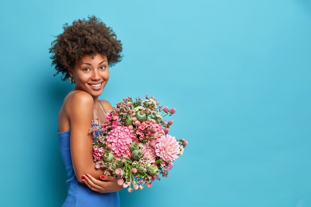 Foto lateral de uma jovem vestindo um vestido segurando um buquê de flores e recebendo como presente em poses de 8 de março contra uma parede azul com espaço de cópia para sua promoção