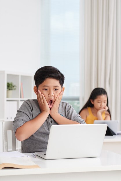 Foto média de alunos asiáticos em sala de aula trabalhando em laptops, um garoto na frente com uma expressão de surpresa no rosto